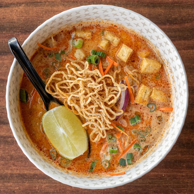 Overhead view of a bowl of Thai Khao Soi with tofu, crispy noodles, and a lime wedge.