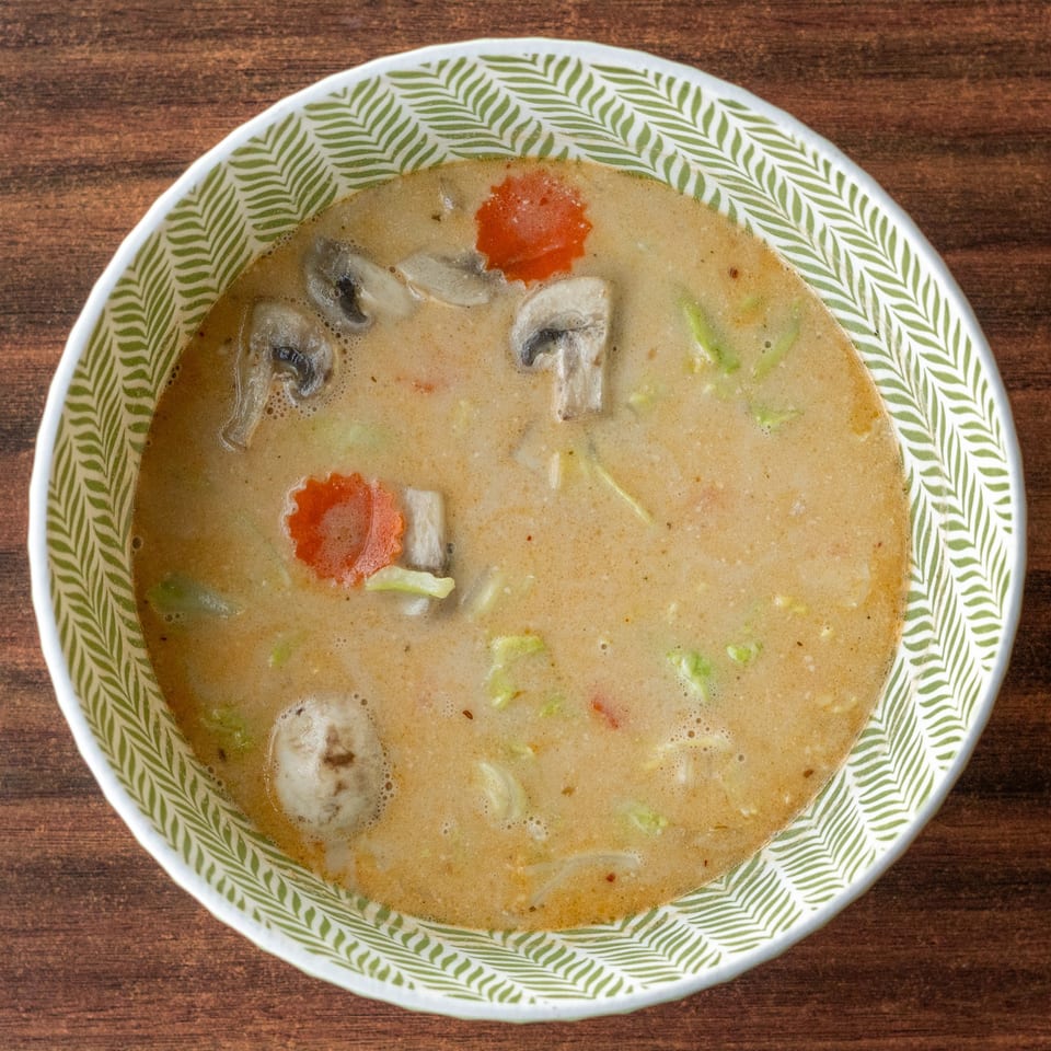 Creamy Tom Kha soup with mushrooms, carrots, and cabbage in a decorative bowl on a wooden table.
