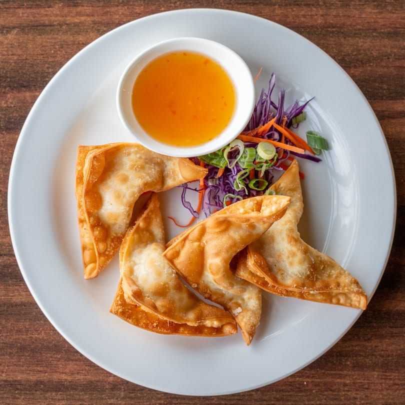 Crispy golden wontons with a side of sweet chili dipping sauce and shredded vegetables on a white plate.