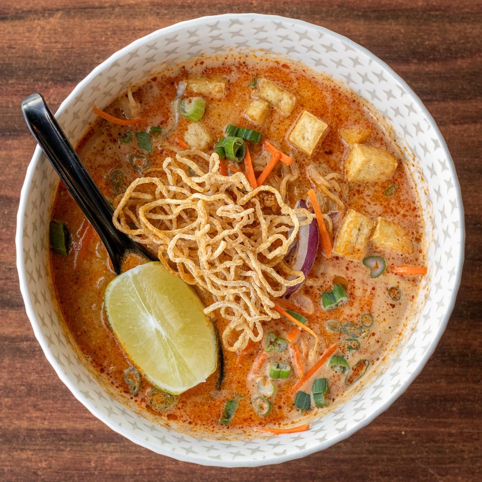 Overhead view of a bowl of curry noodle soup with tofu, lime, and crispy noodles.