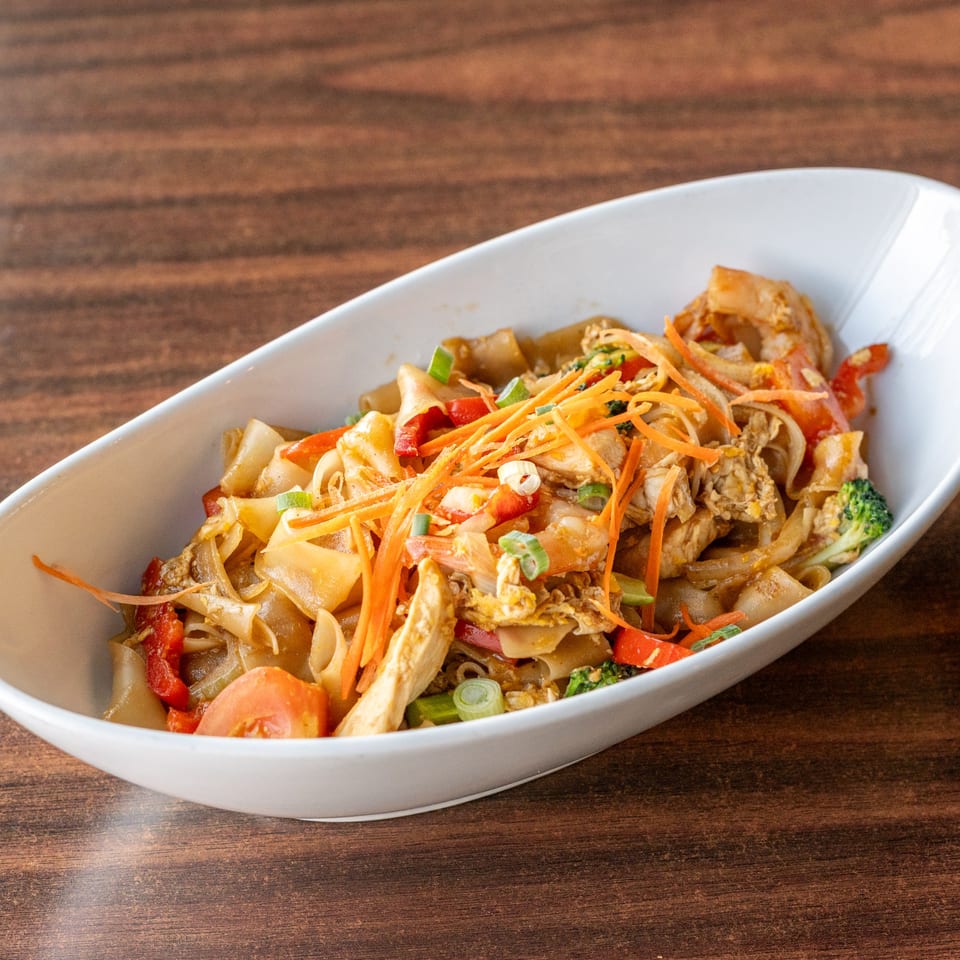 Pad See Ew with chicken, wide noodles, broccoli, carrots, and red peppers in a white bowl.