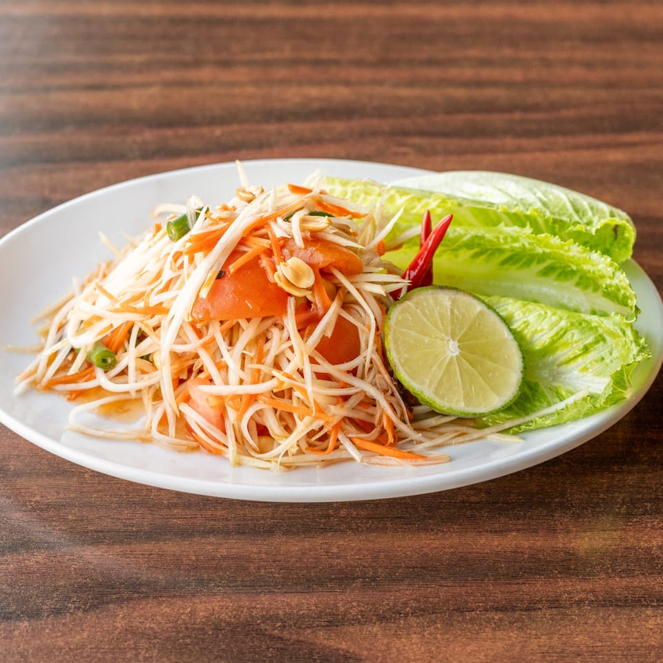 A vibrant papaya salad with shredded carrots, tomatoes, peanuts, lime, and lettuce on a white plate.